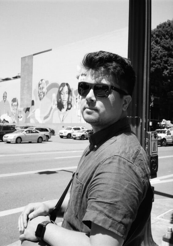 A man wearing sunglasses and a short-sleeved shirt stands on a city street, leaning against a pole. Behind him, a mural featuring various faces covers a large wall. Several cars are parked along the street, and trees are visible in the background.