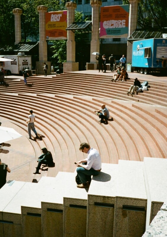 People are scattered across the steps of an outdoor amphitheater, engaging in various activities. Some are sitting and using their phones, while others are walking. Colorful banners and a food stand can be seen in the background, indicating a cultural event.