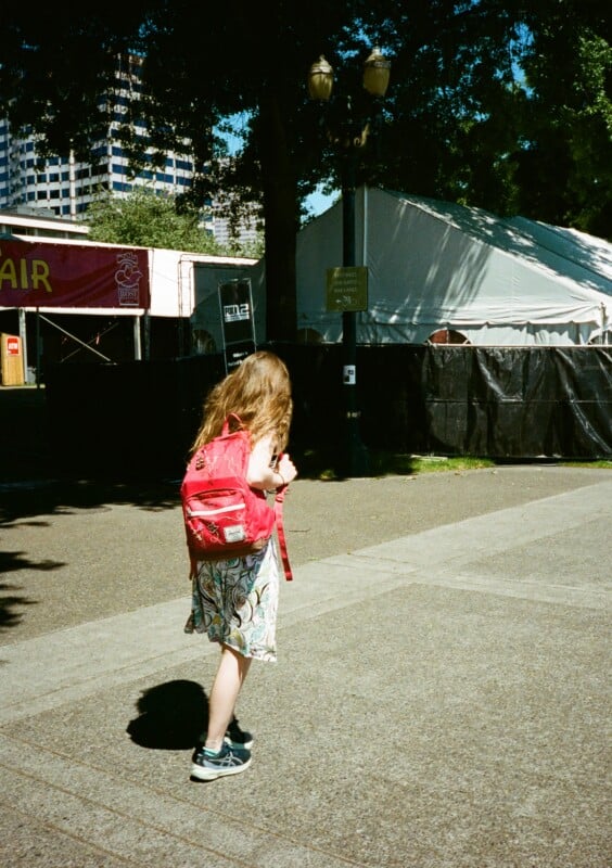 A person with long hair wearing a light summer dress and carrying a pink backpack is walking on a paved walkway. Large tents and multi-story buildings are seen in the background under a clear blue sky.