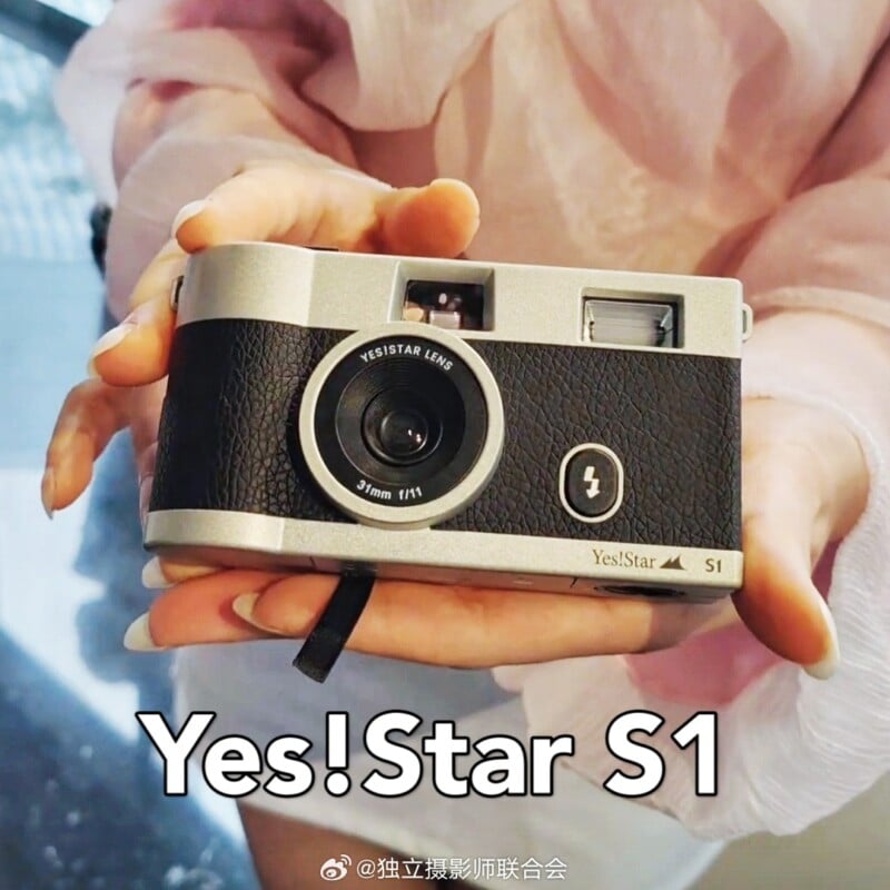 A person holds a compact, retro-style camera labeled "Yes!Star S1." The camera has a black textured grip and a large lens in the center, with "Yes!Star S1" written below. The person's hands are partially visible, wearing a light pink, sheer sleeve.