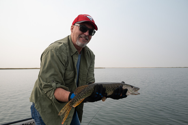 A man wearing a red cap, sunglasses, and a green shirt smiles while holding a fish he caught. He is standing on a boat with a body of water visible in the background under a clear sky.