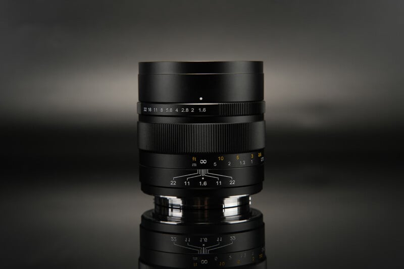 A close-up shot of a black camera lens on a reflective black surface. The lens features white and yellow markings indicating various aperture and focus settings, with a sleek and modern design. The background is a gradient of dark shades.