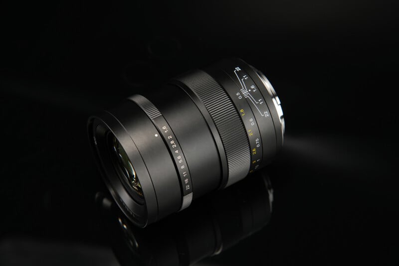 Close-up image of a black camera lens with focus and aperture rings, lying on a reflective black surface. The lens features various markings and numbers indicating different settings. The background is dark, emphasizing the lens's sleek and professional design.