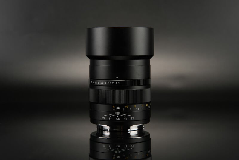 A sleek black camera lens with focus and aperture rings is standing upright against a dark, reflective background. The aperture ranges from f/2.8 to f/22, and various distance markings in feet and meters are visible on the focus ring.