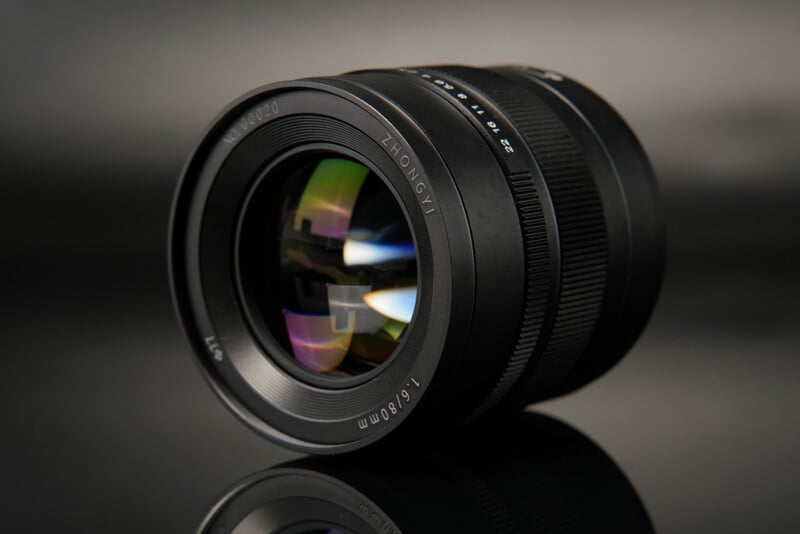 A close-up of a Zhongyi camera lens with "17-50mm" and "1:0.95" markings. The lens is placed on a reflective black surface, capturing vibrant reflections of light on the glass. The background is blurred with a gradient from black to gray.