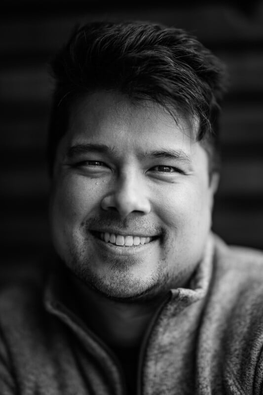 A black-and-white close-up photograph of a man smiling. He has short, tousled hair, light facial hair, and is wearing a zip-up jacket. The background is dark and blurred, drawing focus to his face.