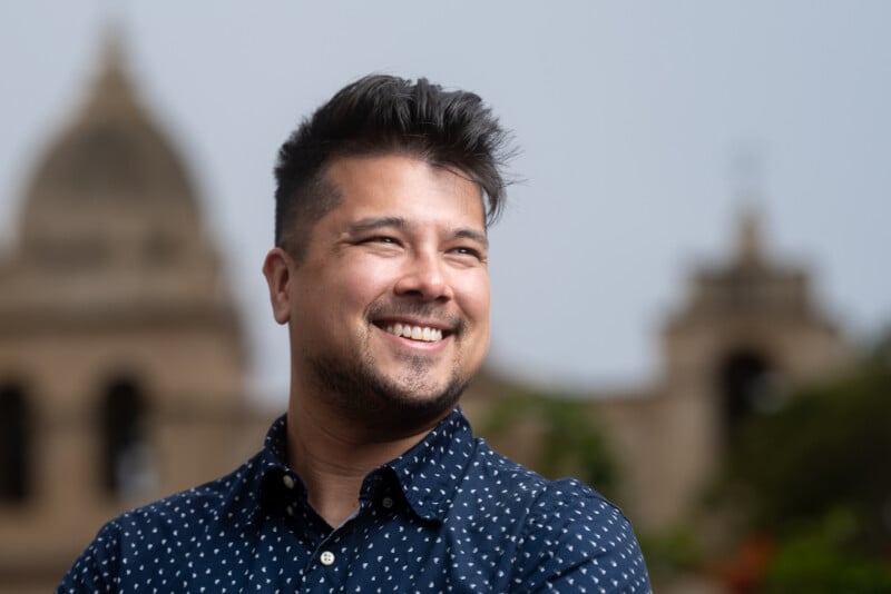 A person with short dark hair and a beard is smiling while looking away from the camera. They are wearing a blue polka-dotted shirt. The background features blurred historic buildings and a cloudy sky.