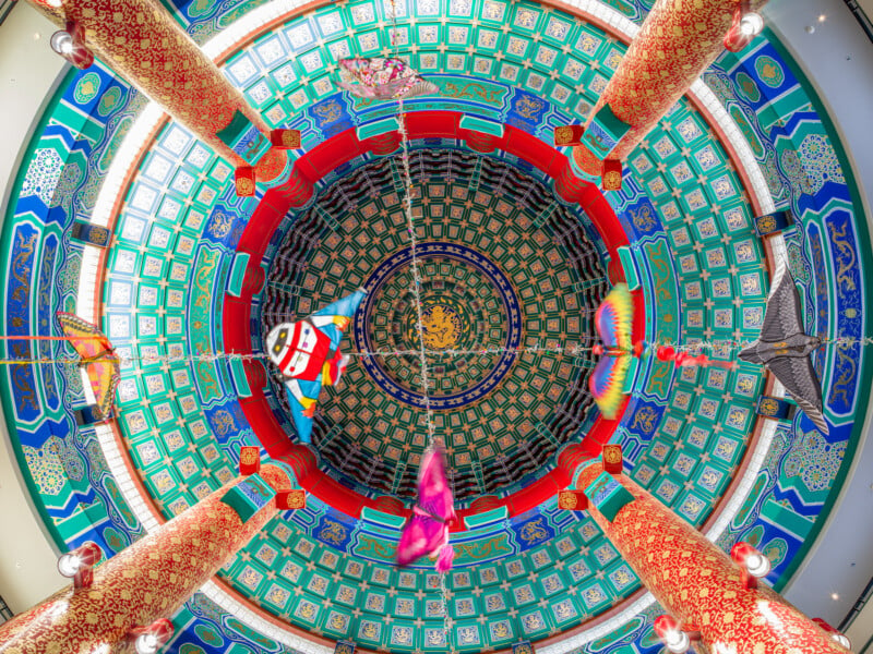 Interior view of a vibrant, ornately decorated temple ceiling showcasing intricate patterns and a central dragon motif, with colorful lanterns and kites hanging below.