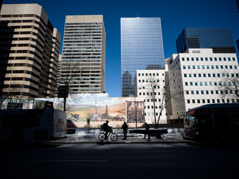 Silhouettes of three people walking and biking in a sunlit urban area, with tall buildings, a mural, and buses under a clear blue sky.