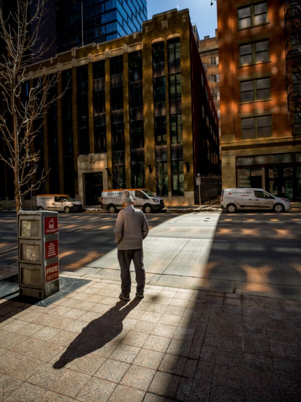 An older man in a jacket and cap stands at a sunny crosswalk in a city, looking towards the shaded street flanked by modern buildings. shadows create a stark contrast on the pavement.