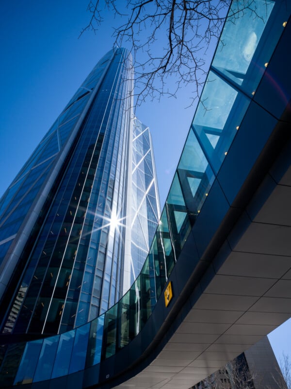 A dynamic view of modern skyscrapers with reflective glass facades under a clear blue sky, featuring a foreground of a curved building with a sunburst reflecting off one tower.