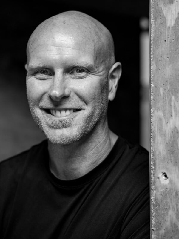 Black and white portrait of a smiling bald man with a beard, wearing a dark shirt, partially obscured by a concrete column on the left.