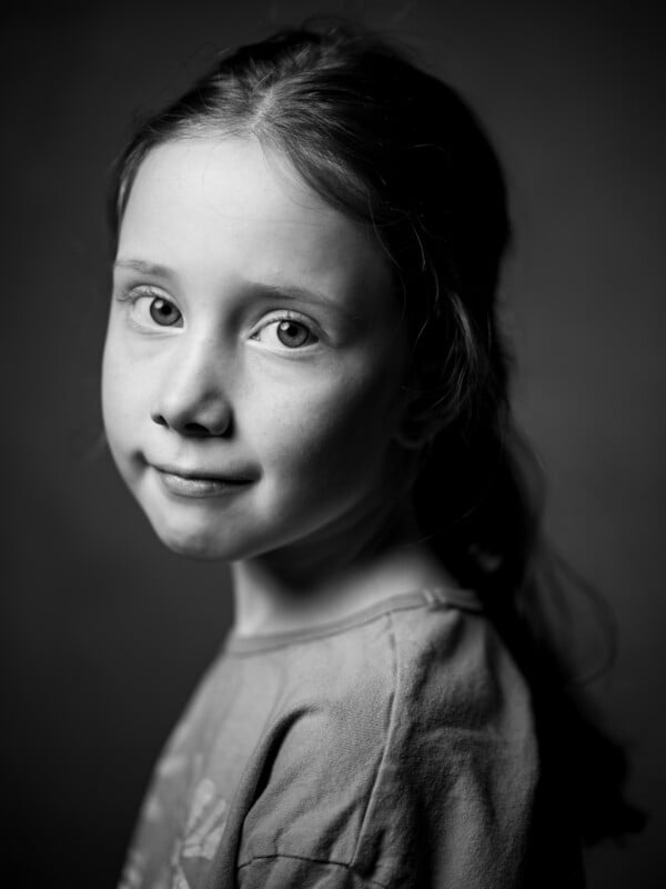 Black and white portrait of a young girl with a subtle smile, looking at the camera. her hair is pulled back and she wears a light blouse. the background is simple and dark, focusing attention on her face.