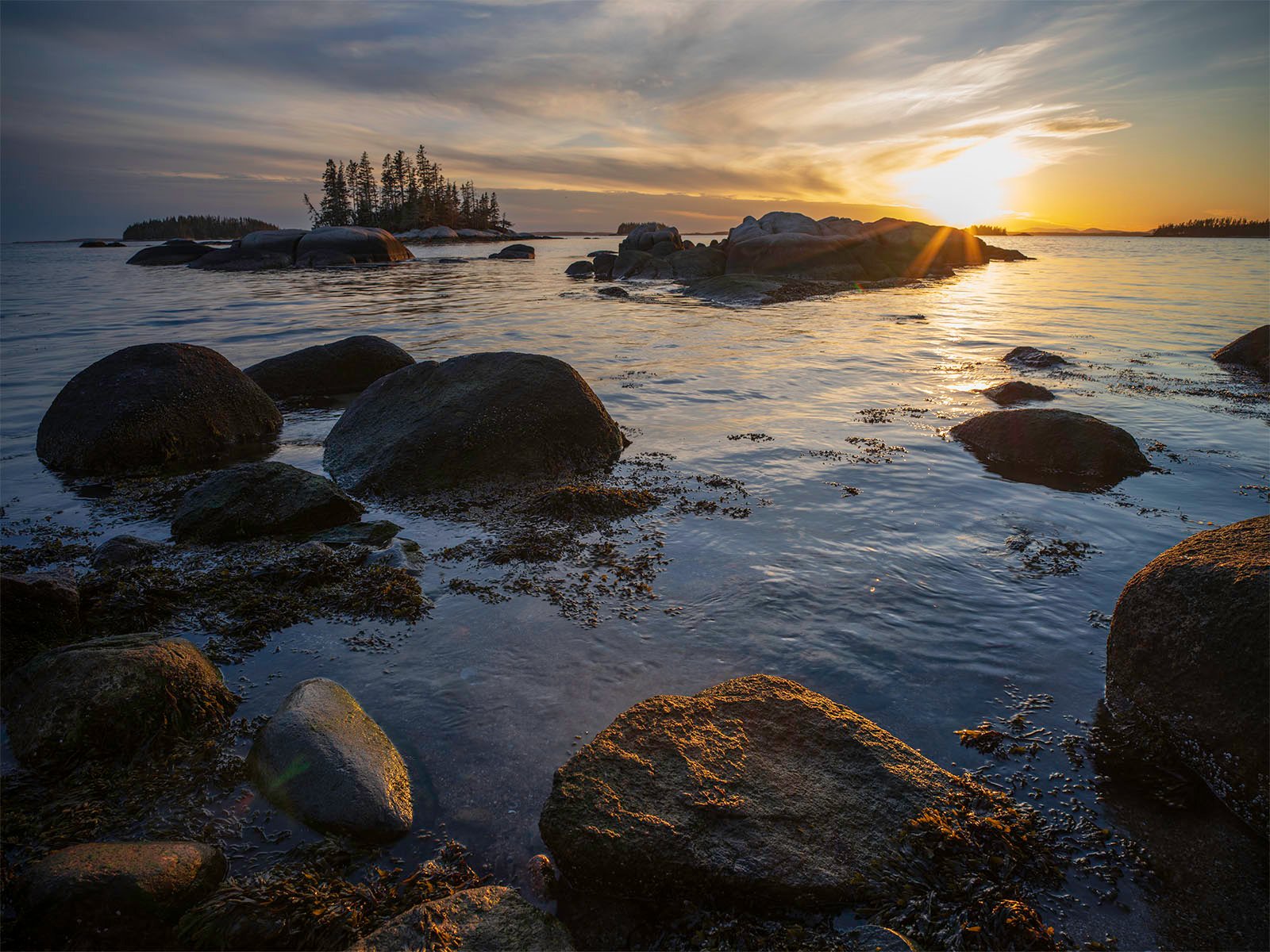 Sunset over a calm sea with rocks in the foreground and small islands in the distance under a golden sky.