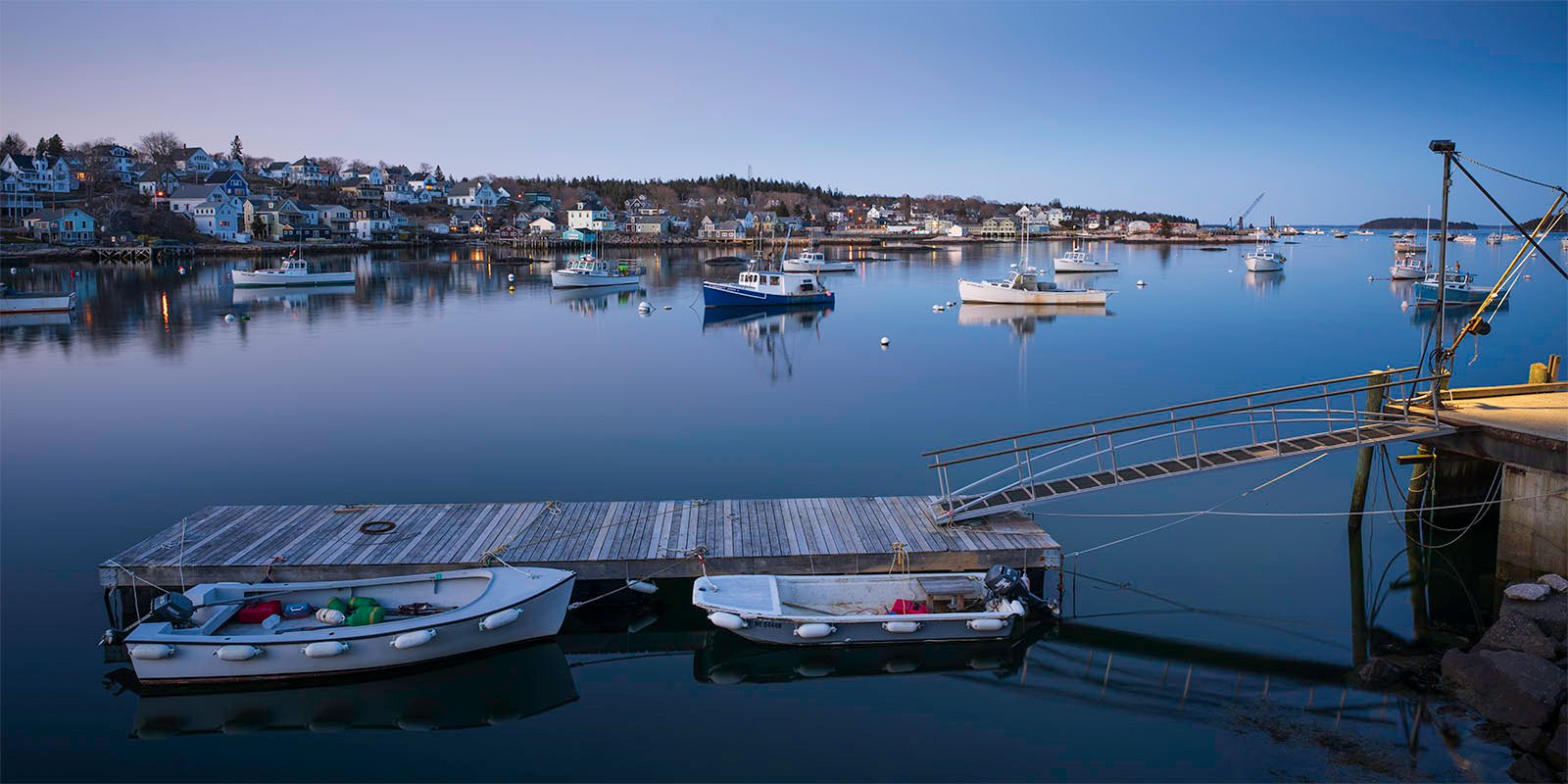 Twilight settles over a serene harbor with boats moored peacefully on calm waters. a small floating dock in the foreground holds two boats, against a backdrop of a quaint hillside town.