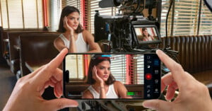 A person in a white tank top with short hair poses inside a diner booth for a video shoot. They rest their head on their hand, looking away. The scene is recorded on a camera, while an individual also views and records it using a smartphone screen in the foreground.