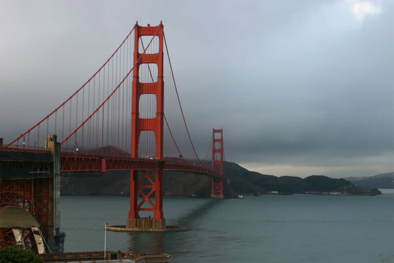 A view of the Golden Gate Bridge spanning across a body of water with overcast skies. The bridge's iconic red-orange towers and cables contrast against the gloomy background. The distant shorelines and hills are faintly visible under the grey clouds.