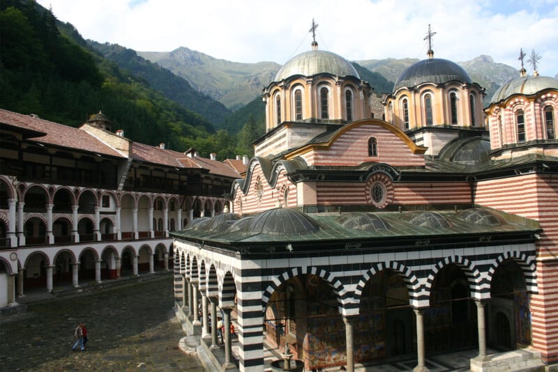 The image shows the Rila Monastery in Bulgaria, with its distinctive striped facade, domed roofs, and a courtyard. Surrounding green mountains evoke a serene atmosphere. A few people can be seen walking in the courtyard, showcasing the monastery's scale and architectural details.