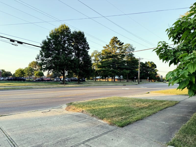 A quiet suburban street scene with a sidewalk in the foreground, a grassy area, and a road with several lanes and power lines overhead. Trees and parked cars are visible in the background. The sky is clear.