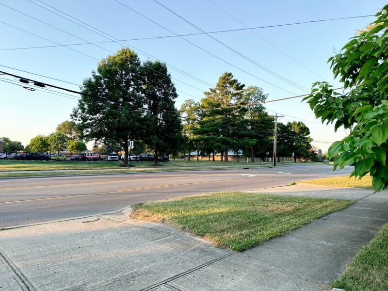 A suburban street with a wide two-lane road lined with grass and sidewalks on either side. Large trees and power lines are visible, and parked cars can be seen in the distance. The sky is clear and the scene is well-lit.