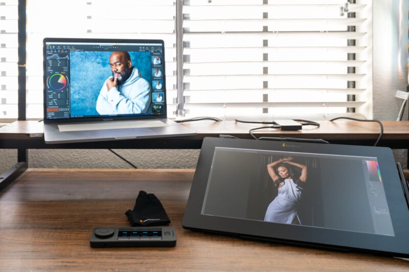 A work desk features a laptop displaying an editing software and a monitor showing a photo of a woman in a white dress. A digital drawing pad and a control device are on the desk. The scene is lit by sunlight through partially open blinds.