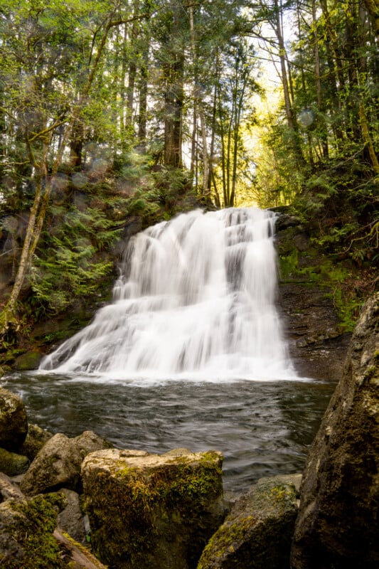 A cascade waterfall surrounded by lush green forests, with sunlight filtering through the trees, shining on the water and moss-covered rocks in the foreground.