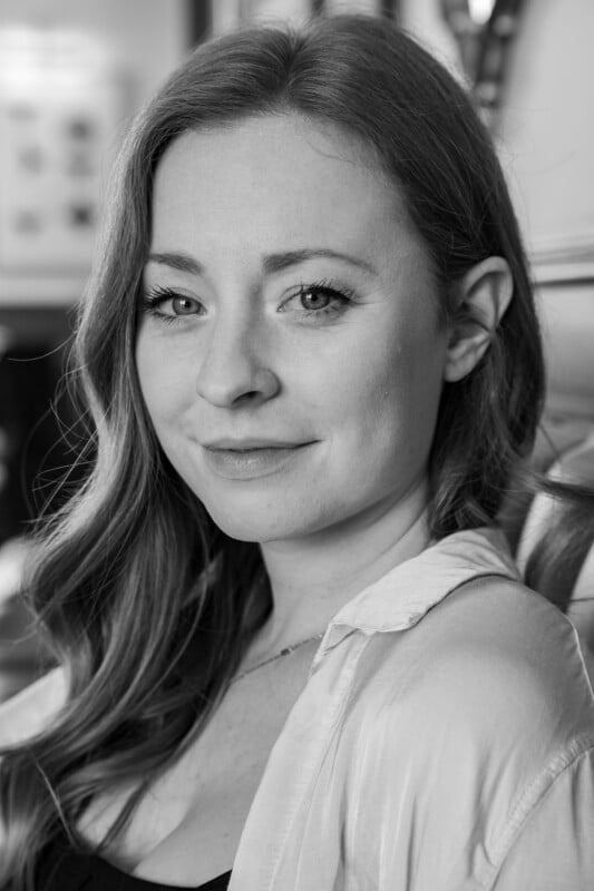 Black and white portrait of a woman with long hair, smiling slightly at the camera. She wears a white shirt and black top, and the background is softly focused.