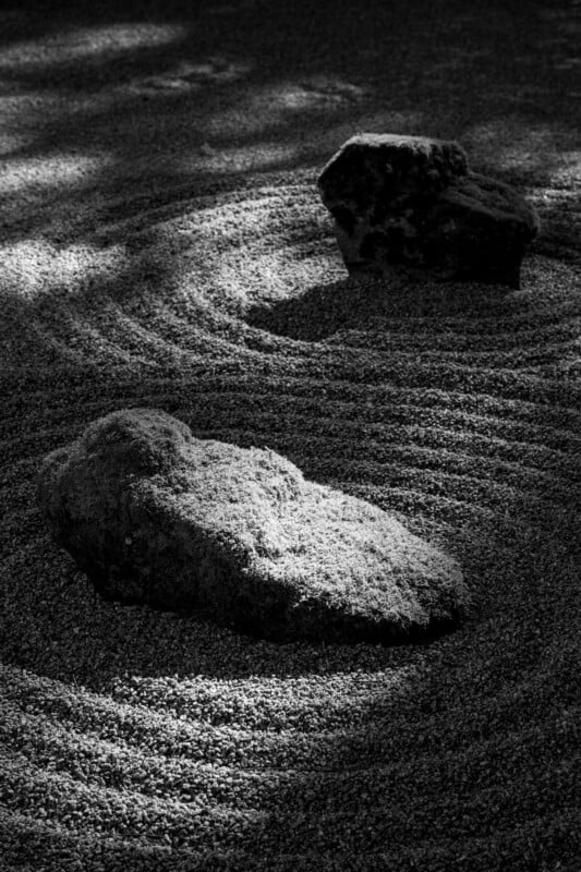 Black and white image of a Japanese rock garden featuring two large, textured rocks. The rocks are placed on gravel meticulously raked into circular patterns around them, creating a serene and harmonious scene. Dappled light and shadows enhance the tranquil atmosphere.