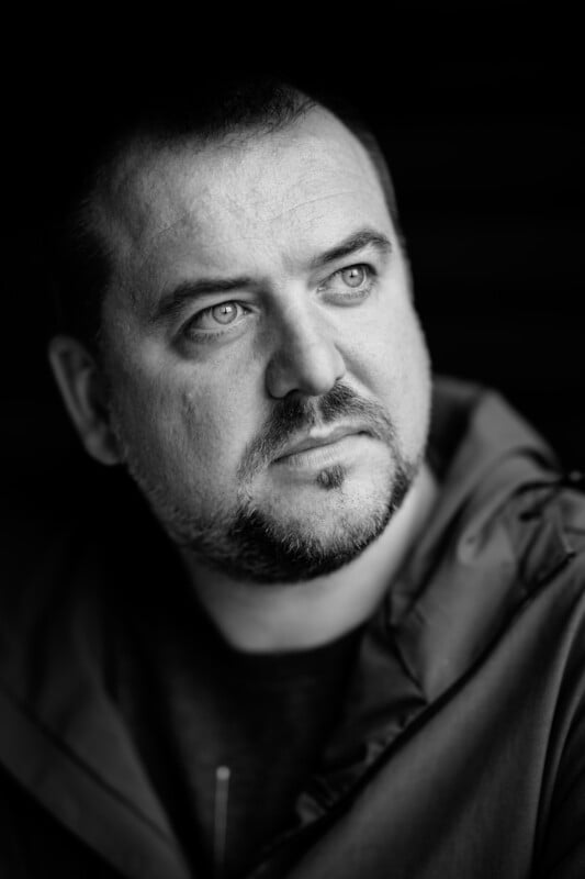 Black-and-white close-up portrait of a man with short hair and a beard. He is looking thoughtfully into the distance. He is wearing a jacket and the background is dark, emphasizing his facial expression.