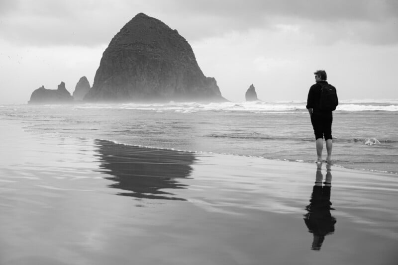 A person stands on a beach, facing away from the camera and looking towards a large rock formation in the ocean. The person's reflection is visible on the wet sand. The sky is overcast, and waves are gently rolling in. The scene is tranquil and contemplative.