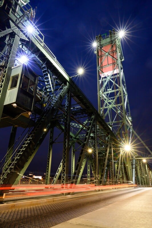 Nighttime view of a lit-up steel truss bridge with multiple lights illuminating the structure. The steel framework and an overhead operator's booth are visible, with light trails from moving vehicles on the bridge. The sky is dark, highlighting the bridge's lighting.