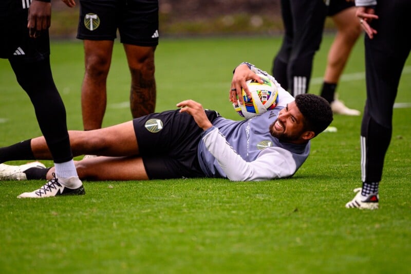 A soccer player wearing a Portland Timbers training kit is lying on the grass, smiling and holding a soccer ball. Two other players with similar kits are standing nearby, their legs visible in the frame. The scene appears to be a training session.