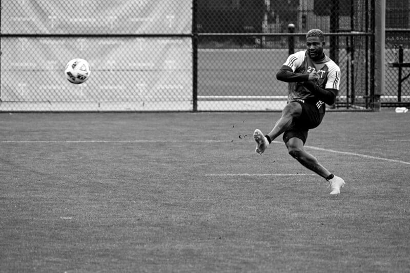 Black and white photo of a soccer player mid-kick on a grassy field. The ball is in mid-air, and the player, wearing athletic gear and cleats, appears focused. A chain-link fence and a partially blurred background suggest the setting is an outdoor practice area.