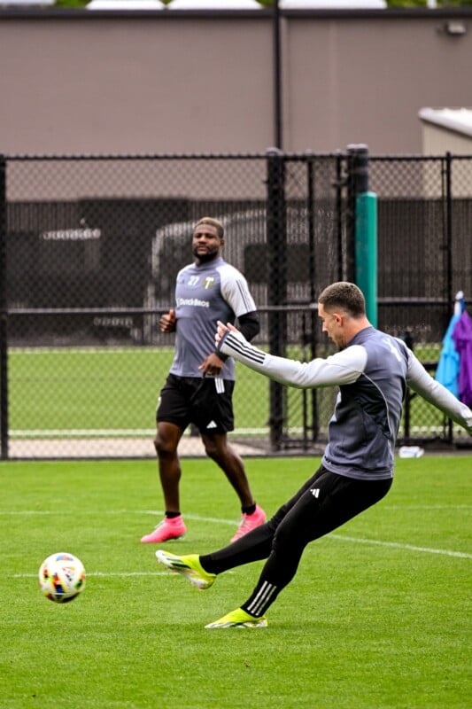 Two soccer players during practice. One in the foreground, wearing a gray top and black pants, is mid-kick, while the other in the background, also in gray, looks on and smiles. They are on a grass field with a chain-link fence in the background. A colorful soccer ball is visible.