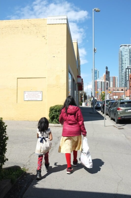 A woman and a child walk down a sidewalk on a sunny day. The woman wears a red jacket, yellow skirt, and red leggings, holding a white shopping bag. The child wears a white top and colorful pants. They are walking by a yellow wall, with buildings visible in the background.