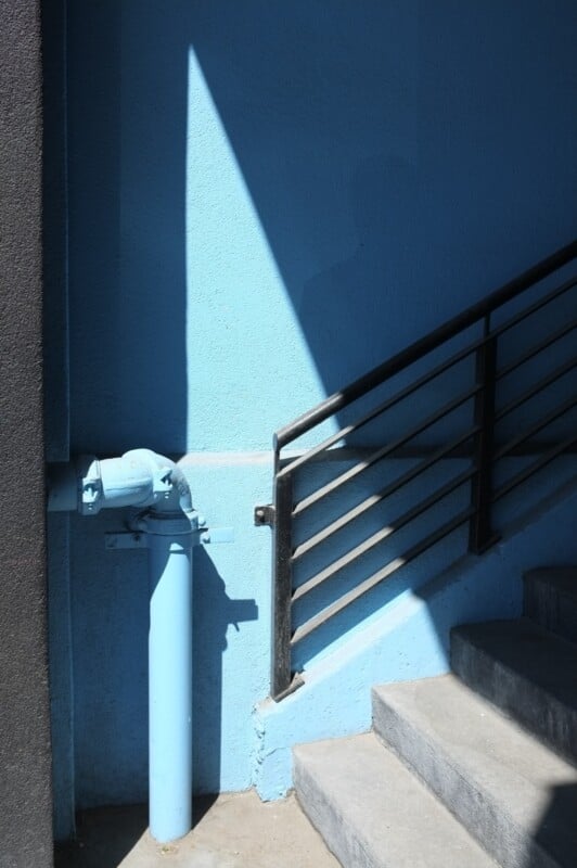 A blue wall with a water pipe attached, painted in the same blue color, is partially shaded. To the right, a handrail follows a concrete staircase ascending diagonally out of the frame. The scene is divided by shadows, creating a stark contrast with the sunlight.