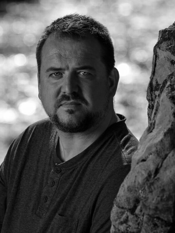 A black and white portrait of a serious-looking bearded man standing next to a textured rock, with bokeh light visible in the background.