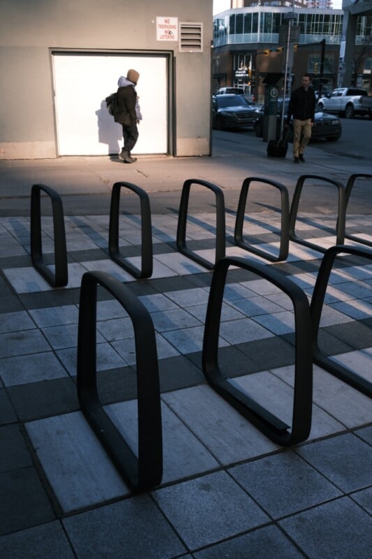 A city street scene with a row of empty bike racks in the foreground. Two people are visible; one walks along the sidewalk near a building with a white door and "No Parking" sign, while the other waits by a busy intersection with cars and taller buildings in the background.