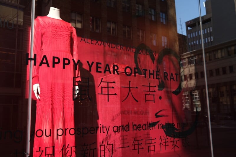 A store display features a red dress on a mannequin with a sign that reads "HAPPY YEAR OF THE RAT" along with Chinese characters. The display also includes a graphic of a stylized rat. The background shows various buildings reflected in the glass.