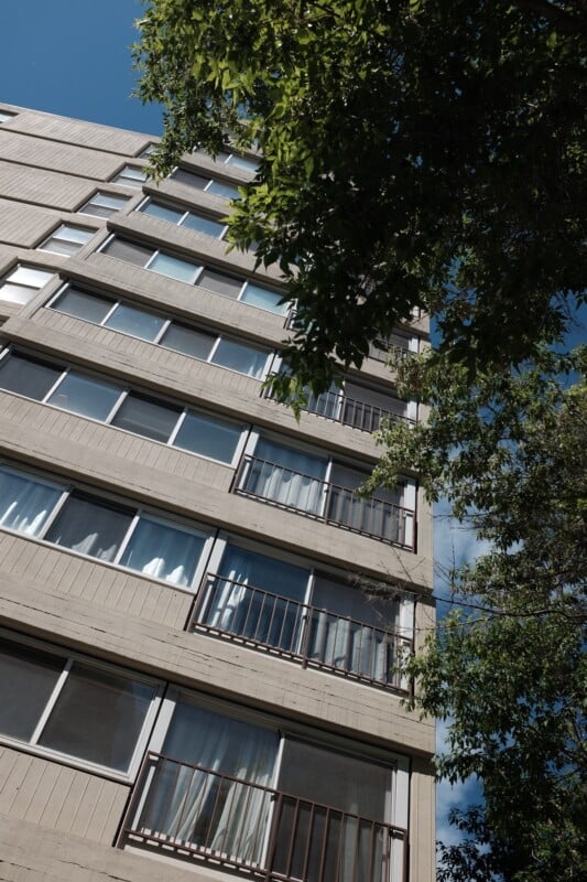 Upward view of a beige apartment building on a clear day. The structure has several floors with windows and small balconies. Green tree branches are visible at the top right, partially covering the building against a bright blue sky.