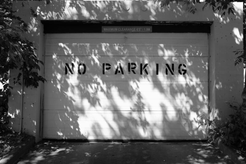 A garage door with the words "NO PARKING" painted on it in all capital letters. Above the door, a sign reads, "MAXIMUM CLEARANCE 6'5" | 1.9M." Dappled sunlight and shadows from nearby trees create a pattern on the garage door and surrounding area.