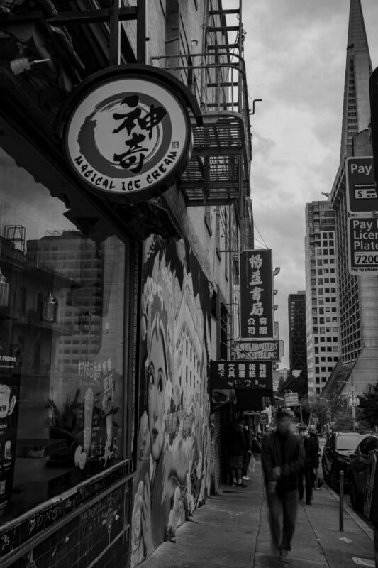 A black and white photograph showing a bustling city street; a storefront with a sign reading "magical ice cream" and artwork of animals on its wall, a blurred pedestrian walking by, and skyscrapers in the background.