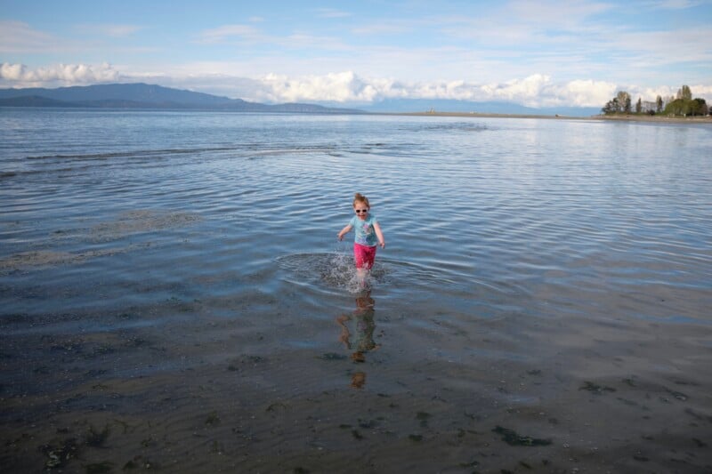 A young child wearing sunglasses, a light blue shirt, and pink pants stands in the shallow waters at a beach. The child is splashing water, and in the background, there are mountains, a cloudy sky, and a tree-lined shoreline.