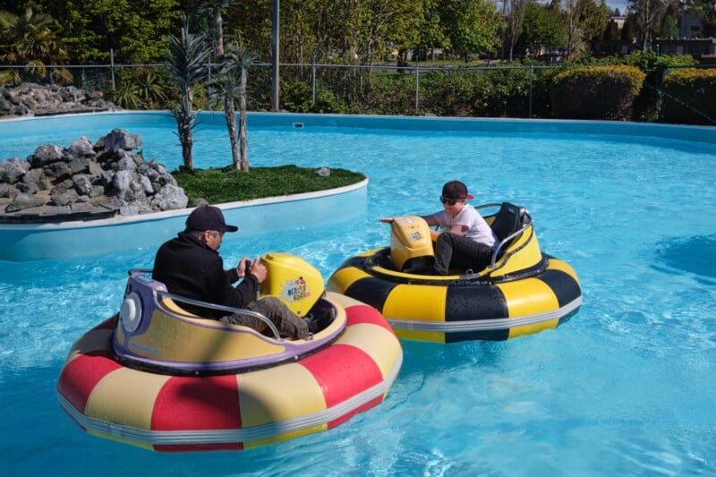 Two people in bumper boats, one wearing a black cap and jacket and the other wearing a white shirt and black cap, are navigating a section of a water park. The scene is set with clear blue water, rocks, and greenery under bright daylight.