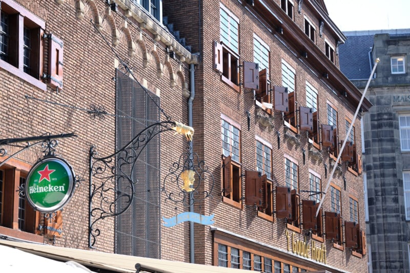 A brick building with many windows featuring wooden shutters in Amsterdam. A Heineken sign hangs from an ornate wrought iron frame on the left side of the building. An awning is partially visible at the lower part of the image.