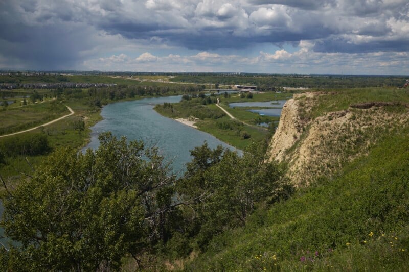 A wide river curves through a lush green landscape with trees, grassy fields, and pathways. A steep cliff covered in grass is on the right, and there are scattered clouds in the sky. A small town or city is visible in the distance.