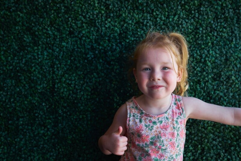 A young girl with light hair pulled back in a ponytail gives a thumbs-up while standing against a background of dense green foliage. She is wearing a sleeveless floral dress and has a cheerful expression on her face.