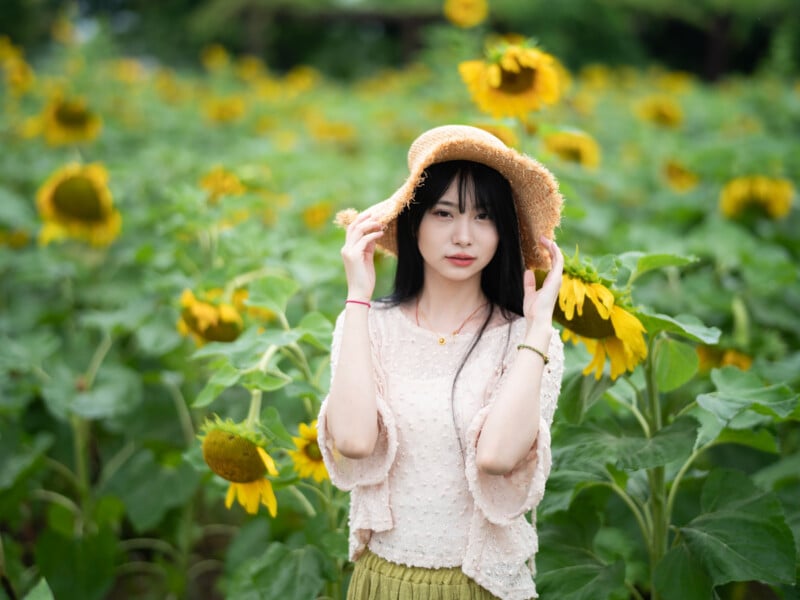 A woman stands in a sunflower field wearing a straw hat, a light pink blouse, and a green skirt. She is holding the brim of her hat with both hands and is looking directly at the camera. Sunflowers surround her, creating a vibrant and cheerful backdrop.