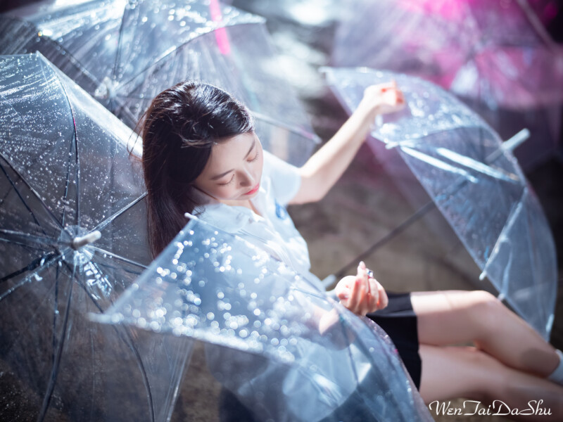 A woman with long dark hair is sitting among several transparent umbrellas on a rainy day. She is wearing a white shirt and a thoughtful expression, with droplets of water on the umbrellas reflecting light around her. The background is blurred.