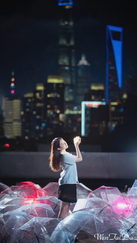 A woman stands amid many umbrellas on a rooftop at night, gazing up with her hands clasped. The city skyline, featuring numerous illuminated buildings, is visible in the background. Soft lighting highlights her and the umbrellas, creating a serene scene.